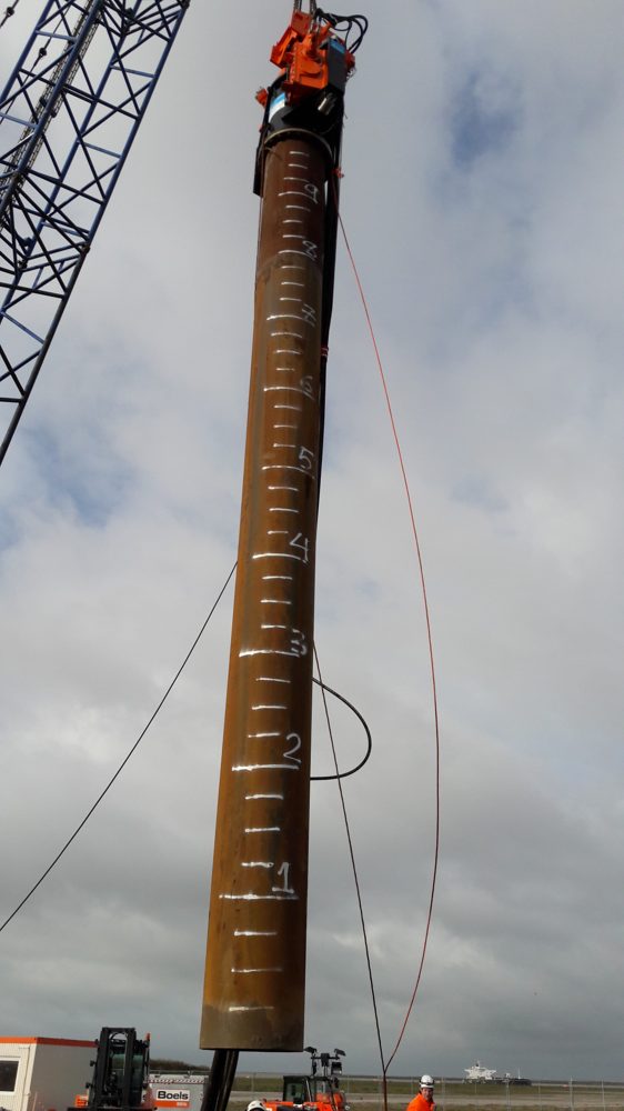 Trilblok bovenop een monopile met dieptemarkeringen, hangend aan een kraan